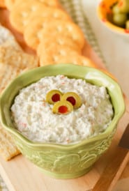 Small bowl holding Green Olive Cheese Spread, with some on a cracker in front and more crackers in back
