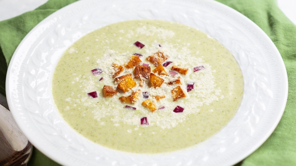close up of white bowl holding one serving of Healthy Broccoli Soup with potato croutons, red onion and Parmesan cheese garnishes