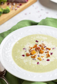 white bowl holding one serving of Healthy Broccoli Soup with potato croutons, red onion and Parmesan cheese garnishes. broccoli florets and partially chopped red onion in the background