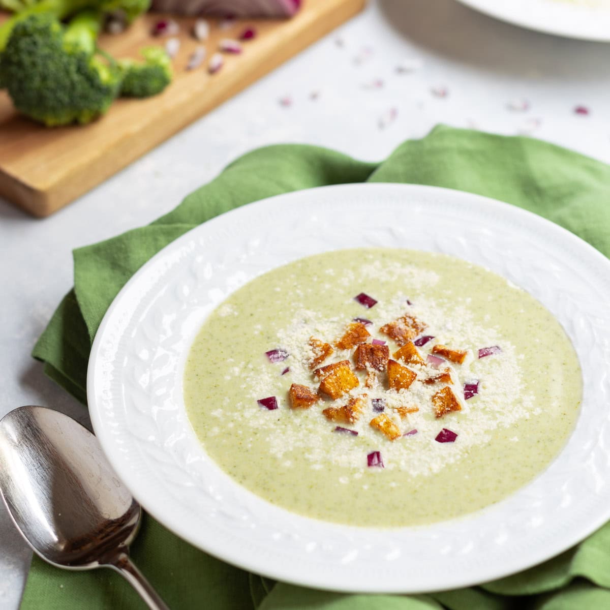 white bowl holding one serving of Healthy Broccoli Soup with potato croutons, red onion and Parmesan cheese garnishes. broccoli florets and partially chopped red onion in the background