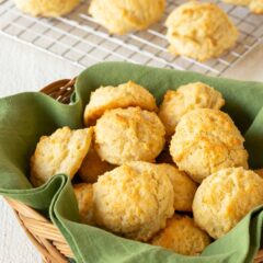 Wicker basket lined with a green napkin and filled with Easy Drop Biscuits, with more biscuits on a cooling rack in the back