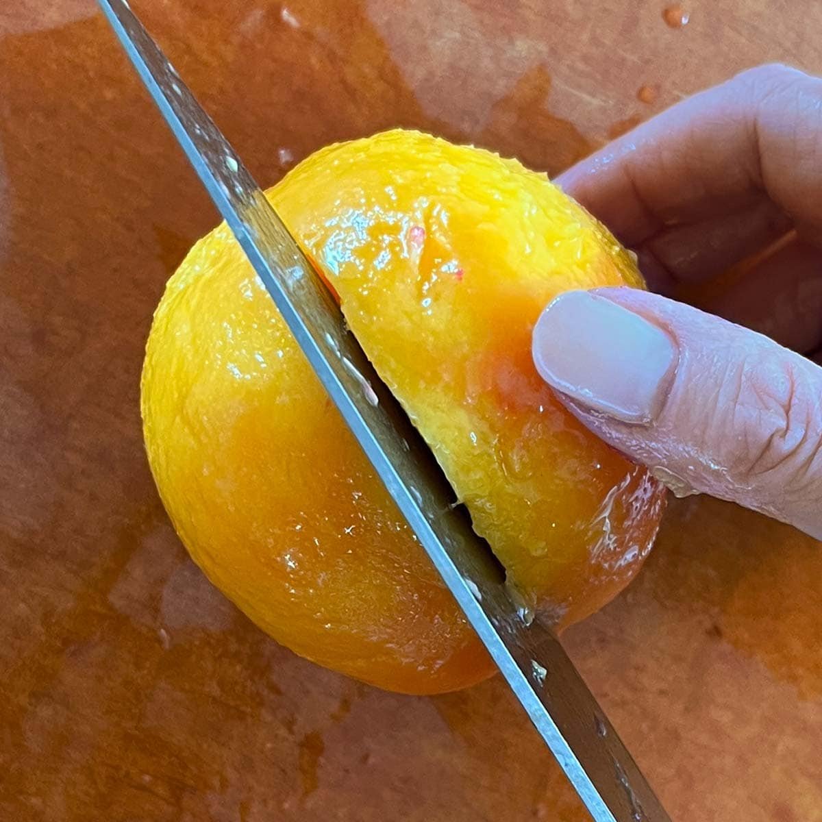 Hand holding peeled peach in place and a sharp knife about halving the peach.
