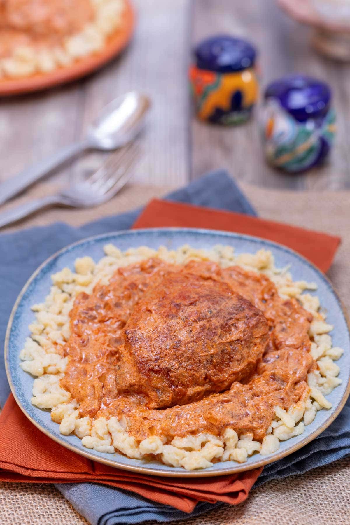 A blue plate holding one portion of Hungarian Chicken Paprikash and dumplighs, with colorful salt and pper in the background