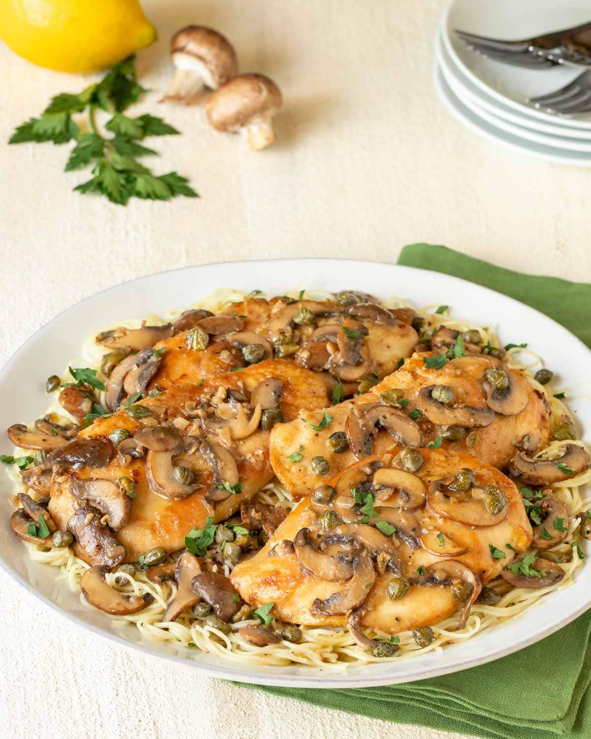 Round white platter holding Lemon Chicken Piccata with Mushrooms atop angel hair pasta, with a fork on the side, a napkin underneath, and lemon and 2 mushrooms in the backgroud with plates