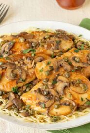 Round white platter holding Lemon Chicken Piccata with Mushrooms atop angel hair pasta, with a fork on the side and a napkin underneath