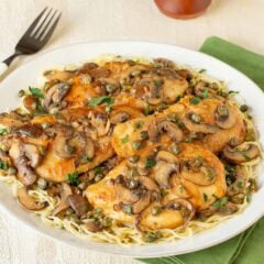Round white platter holding Lemon Chicken Piccata with Mushrooms atop angel hair pasta, with a fork on the side and a napkin underneath