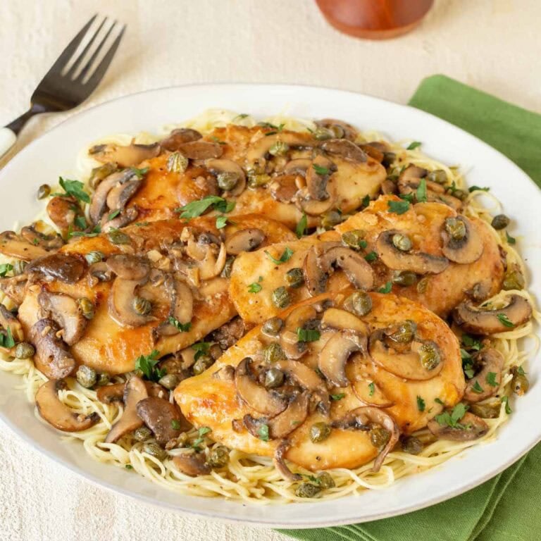 Round white platter holding Lemon Chicken Piccata with Mushrooms atop angel hair pasta, with a fork on the side and a napkin underneath