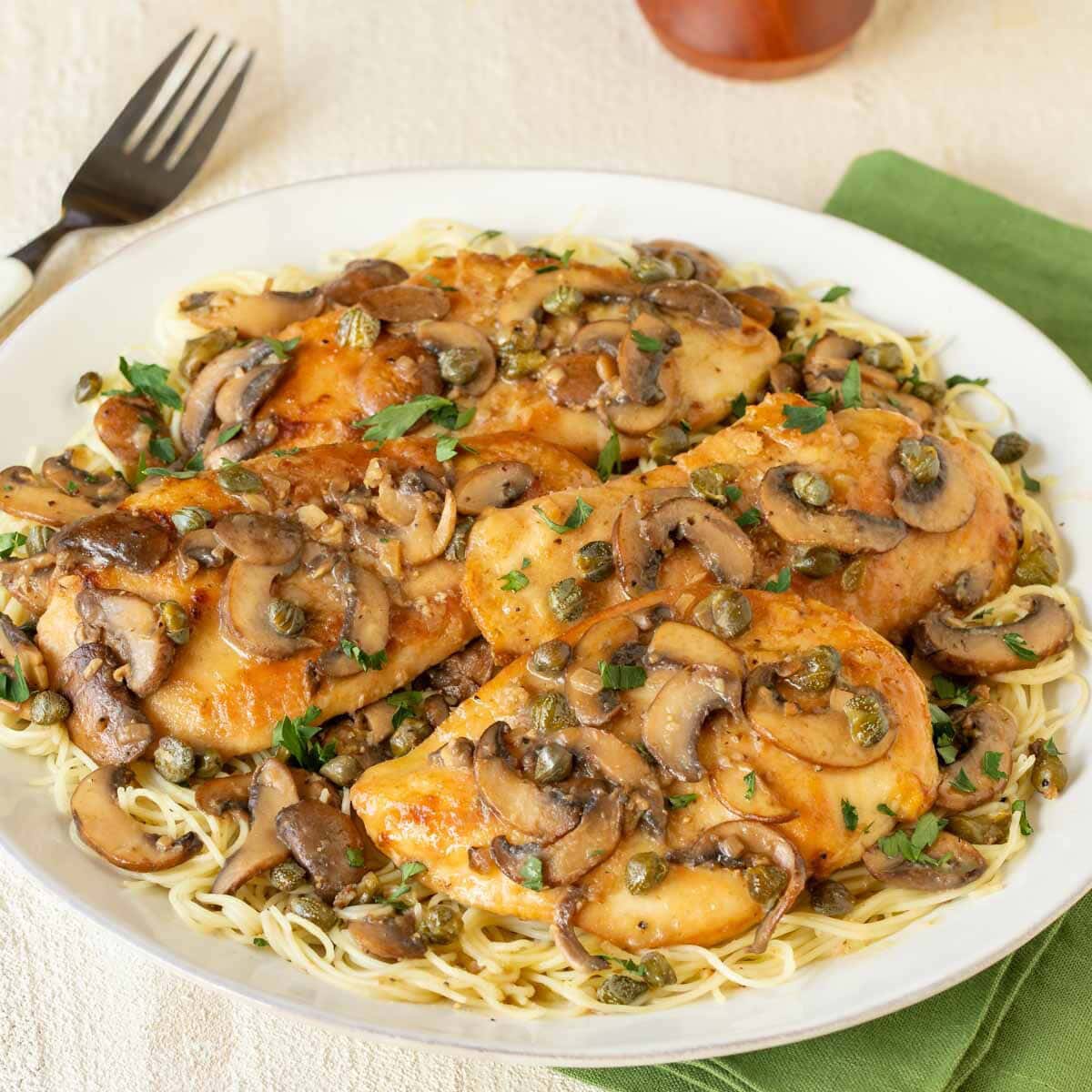 Round white platter holding Lemon Chicken Piccata with Mushrooms atop angel hair pasta, with a fork on the side and a napkin underneath