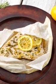 Swordfish fillet topped with lemon slices, lemon zest and chopped rosemary in a parhment paper packge.
