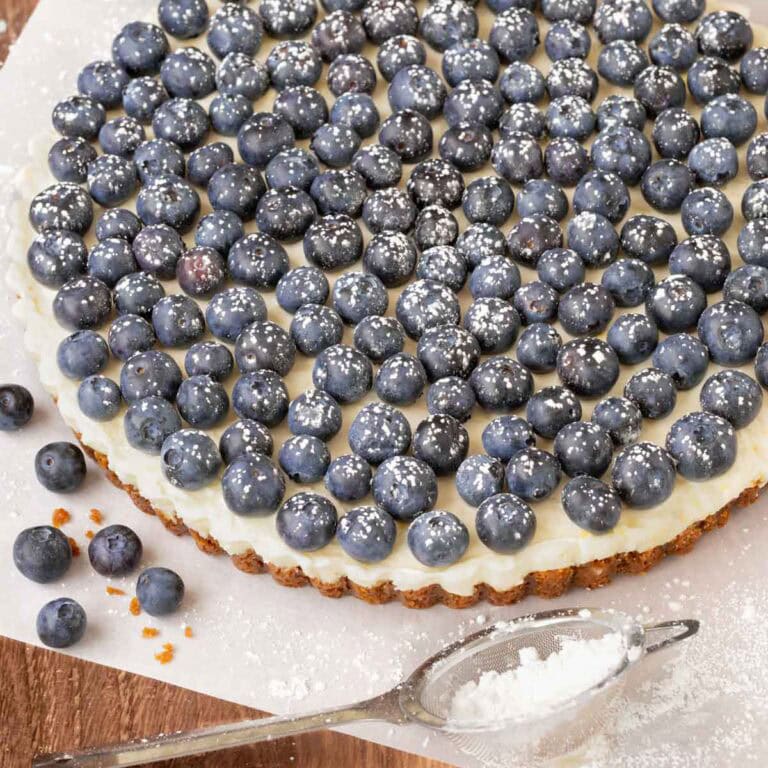 Close up of finished Lemon Tart topped with fresh blueberries and dusted with powder sugar, with a small bowl holding more berries in back and a sifter in front