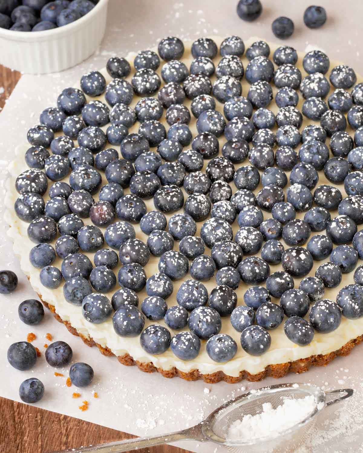 Finished Lemon Tart topped with fresh blueberries and dusted with powder sugar, with a small bowl holding more berries in back and a sifter in front