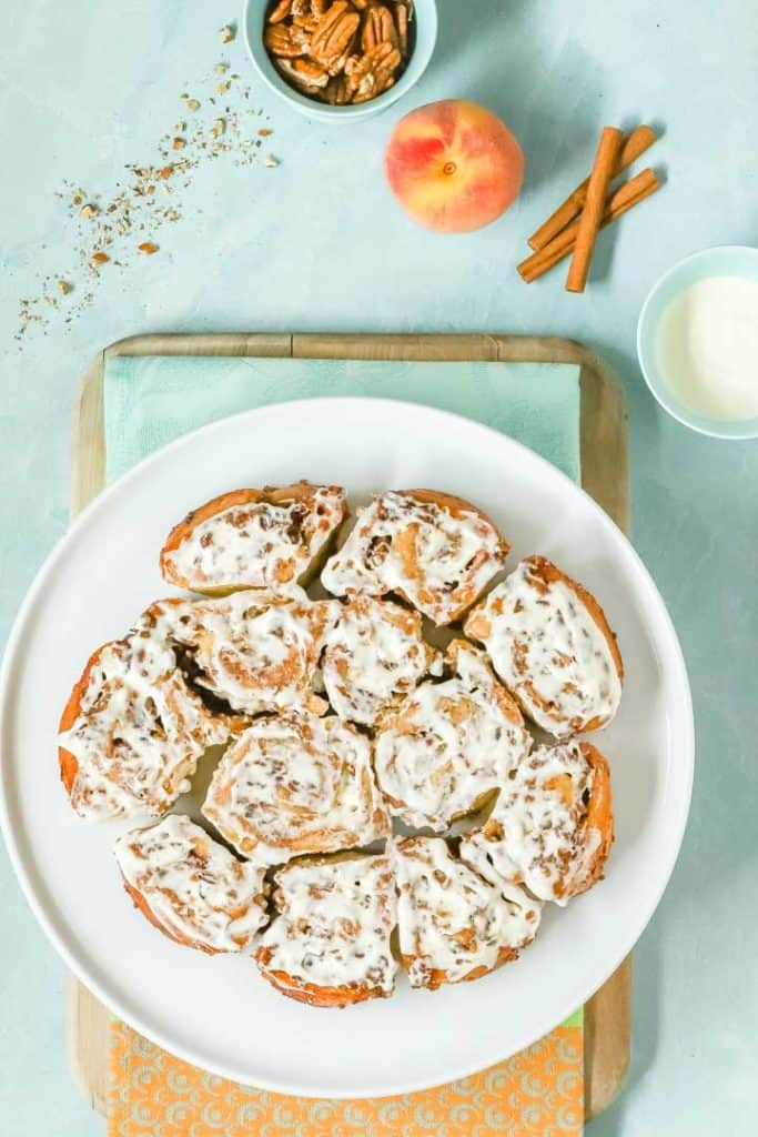 Overhead view of a plate holding a batch of No Yeast CInnamon Rolls with a peach, cinnamon sticks and cream cheese glaze on the side