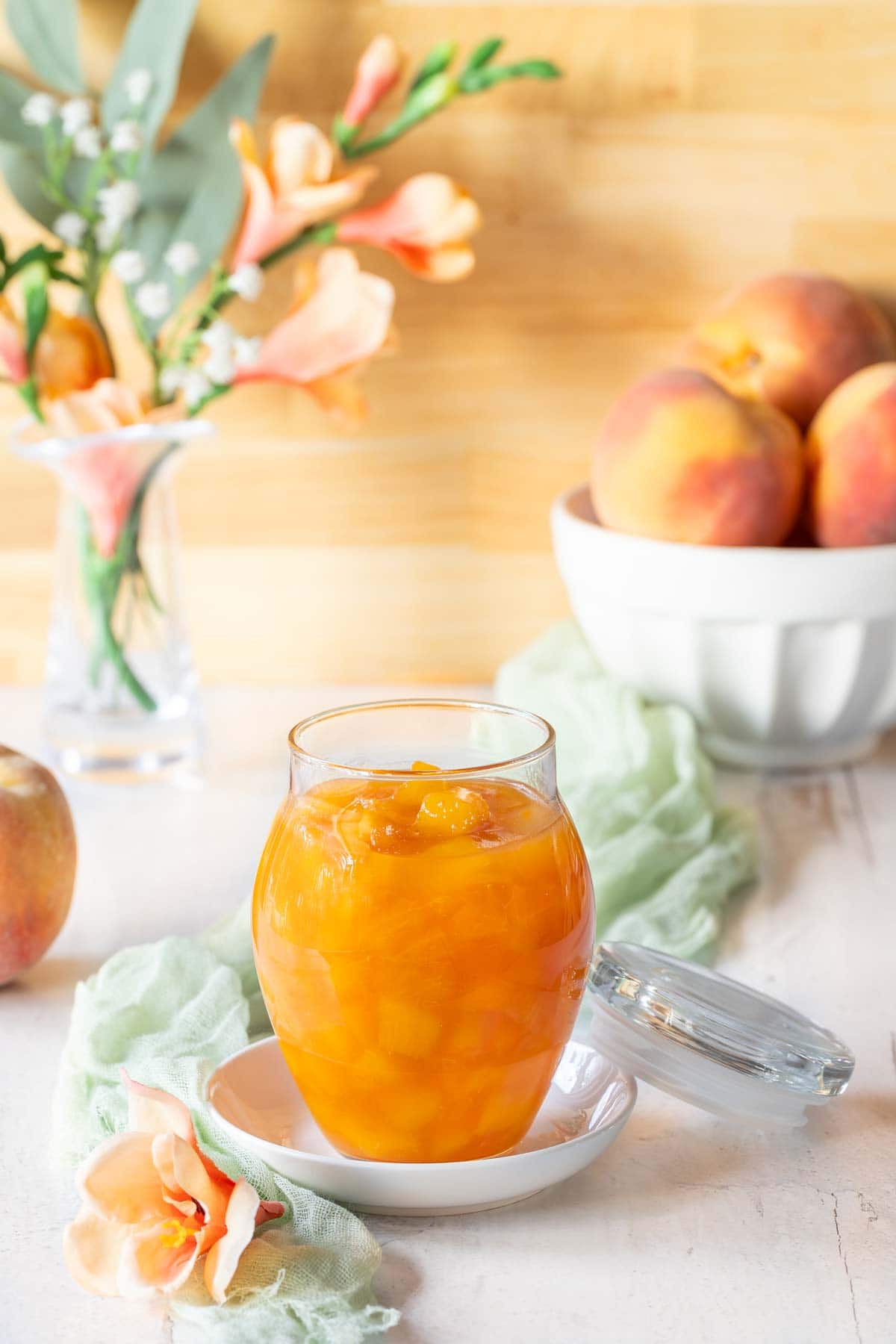 Glass jar holding Peach Sauce, with a bowl of peaches and flowers in the background