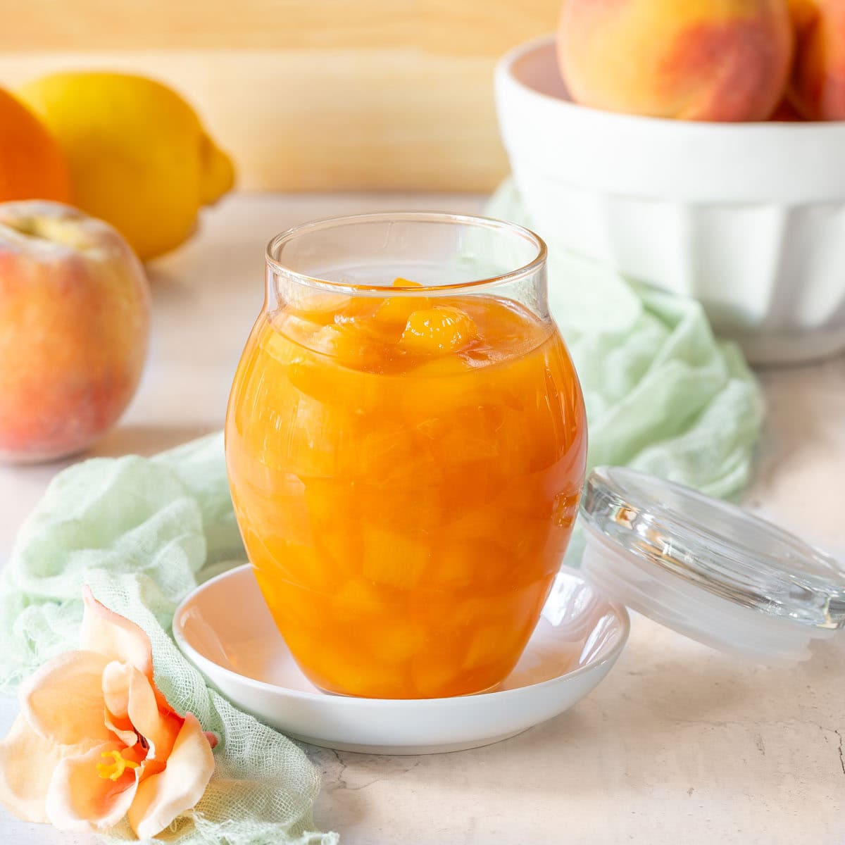 Close up of a glass jar holding Peach Sauce, with a bowl of peaches and a peach, orange, and lemon in the background