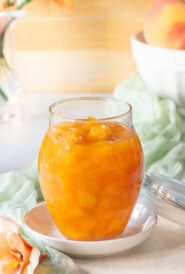 Closer view of a glass jar holding Peach Sauce, with a bowl of peaches and flowers in the background