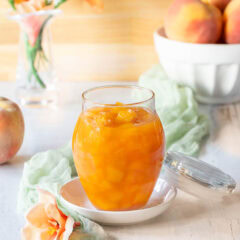 Closer view of a glass jar holding Peach Sauce, with a bowl of peaches and flowers in the background