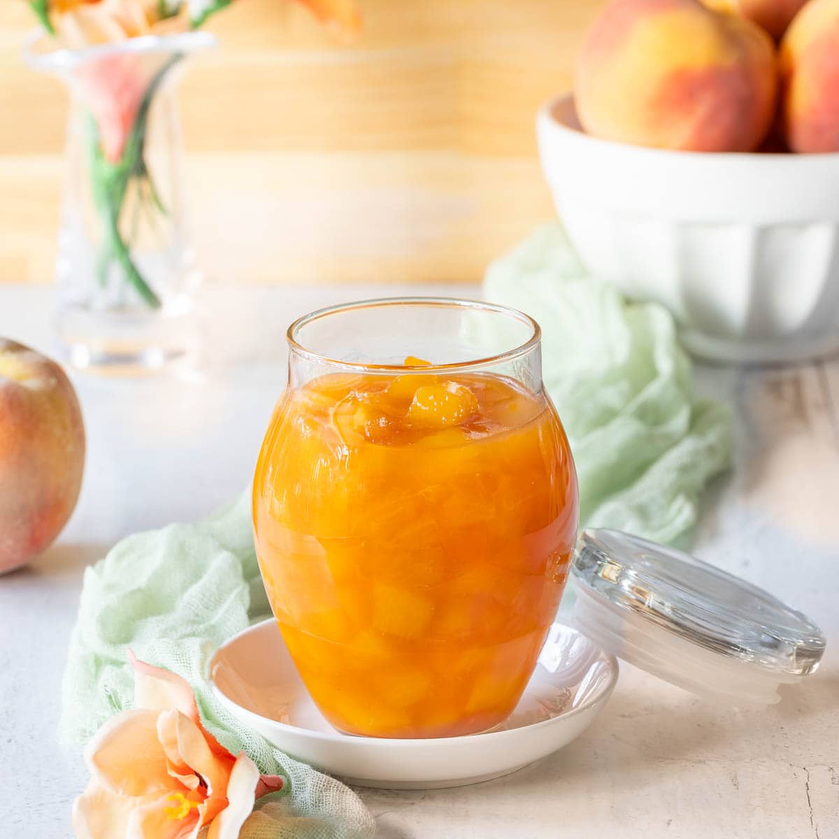 Closer view of a glass jar holding Peach Sauce, with a bowl of peaches and flowers in the background