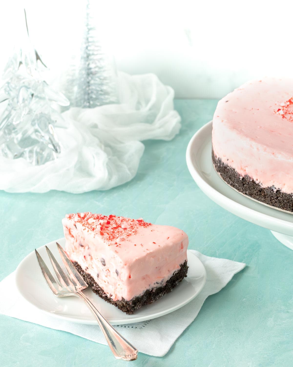 Slice of Frozen Peppermint Chocolate Cheesecake on a white plate, with the rest of the cake in the background alongside glass trees