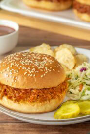 a white plate holding one pulled pork sandwich with BBQ sauce, and with coleslaw chips and pickle slices on the side. A bowl of more BBQ sauce is behind