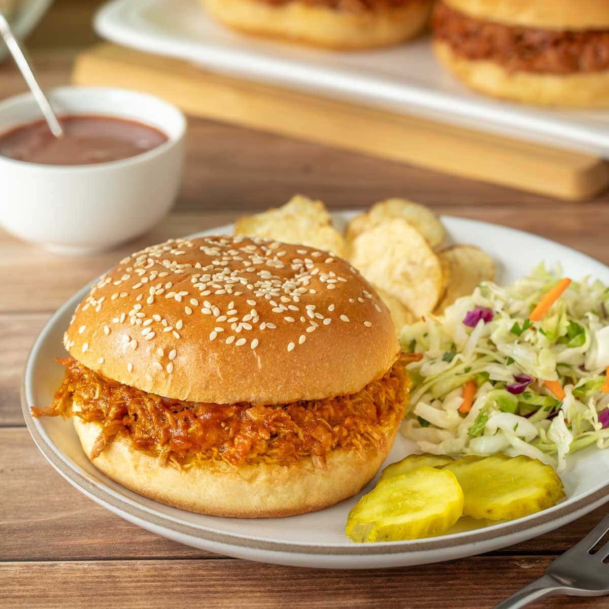 a white plate holding one pulled pork sandwich with BBQ sauce, and with coleslaw chips and pickle slices on the side. A bowl of more BBQ sauce is behind