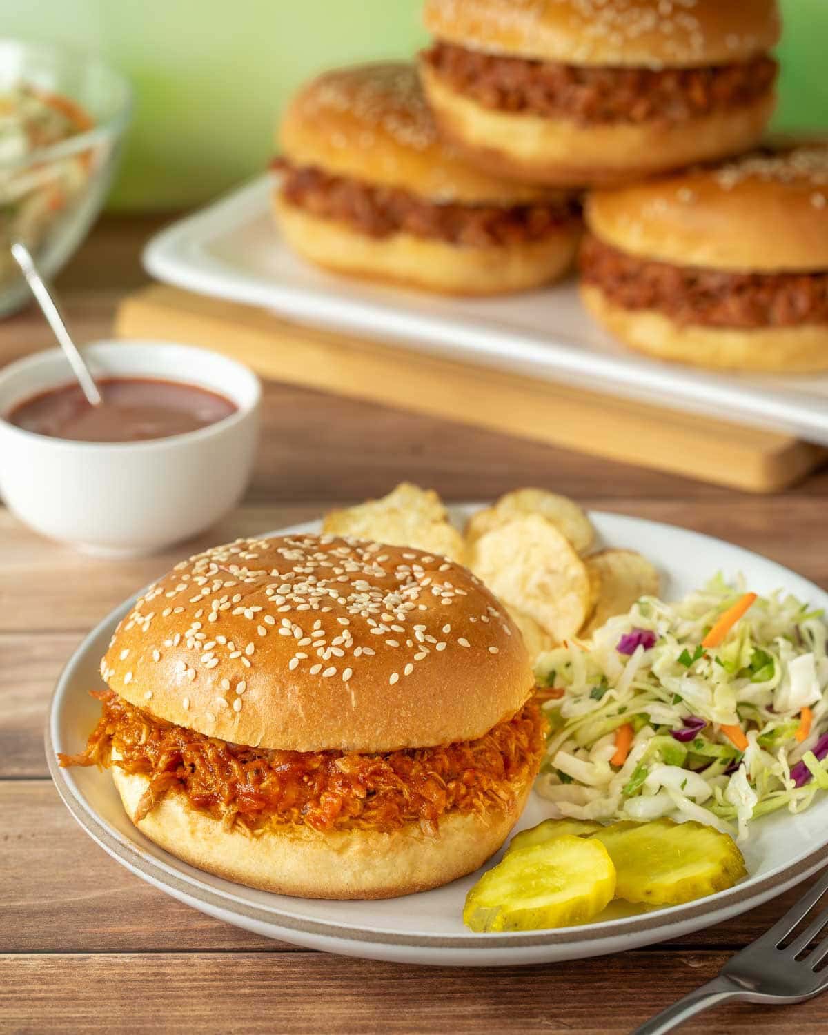 white plate holding one pulled pork sandwich with BBQ sauce, and with coleslaw chips and pickle slices on the side. A plater holds 3 more sandwiches in the background