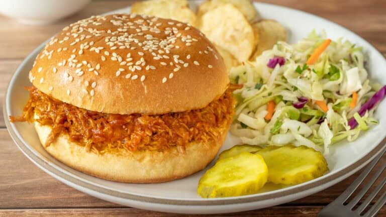 close up of a white plate holding one pulled pork sandwich with BBQ sauce, and with coleslaw chips and pickle slices on the side