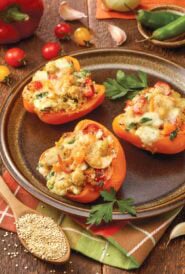 Three Stuffed Bell Peppers with Quinoa on a brown plate, with more ingredients surrounding the plate
