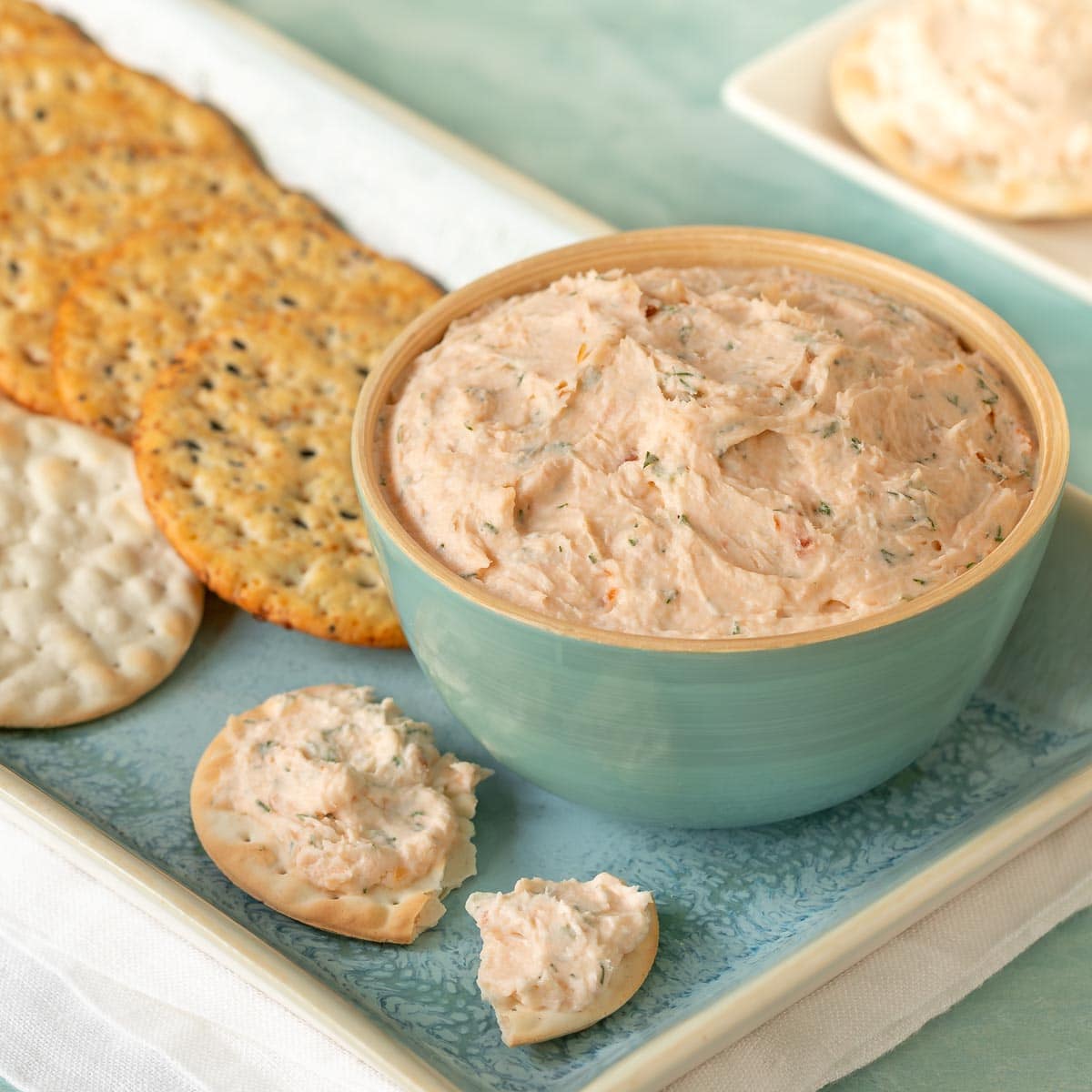 Close up shot of Smoked Salmon Mousse in a blue bowl, with a cracker with mousse on it to the side and more crackers in the back
