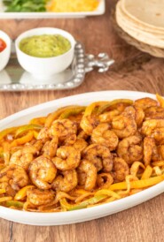 White oval platter holding shrimp fajitas with various toppings and tortillas in the background