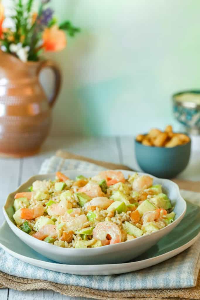 Blue plate holding Rice, Shrimp and Avocado Salad. Bowl of cashews and a pitcher of flowers in the background