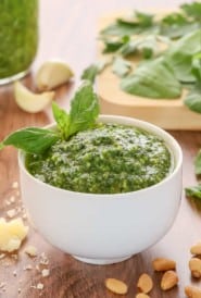 white bowl holding spinachh basil pesto, garnished with a basil leaf and with spinach and more pesto in the background