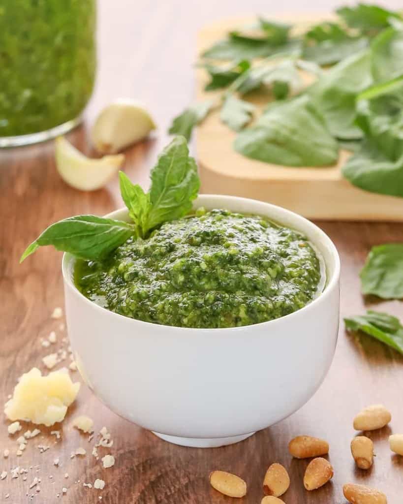white bowl holding spinach basil pesto, garnished with a basil leaf and with spinach and more pesto in the background