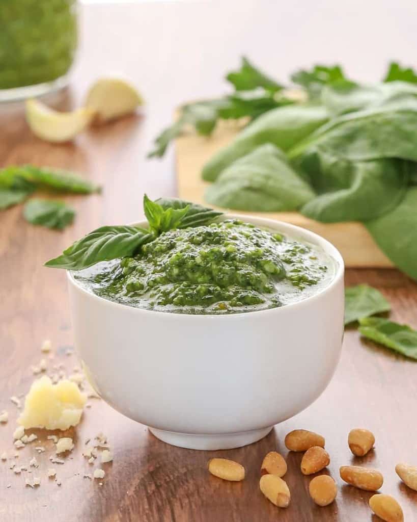 white bowl holding spinach basil pesto, garnished with a basil leaf and with spinach and more pesto in the background