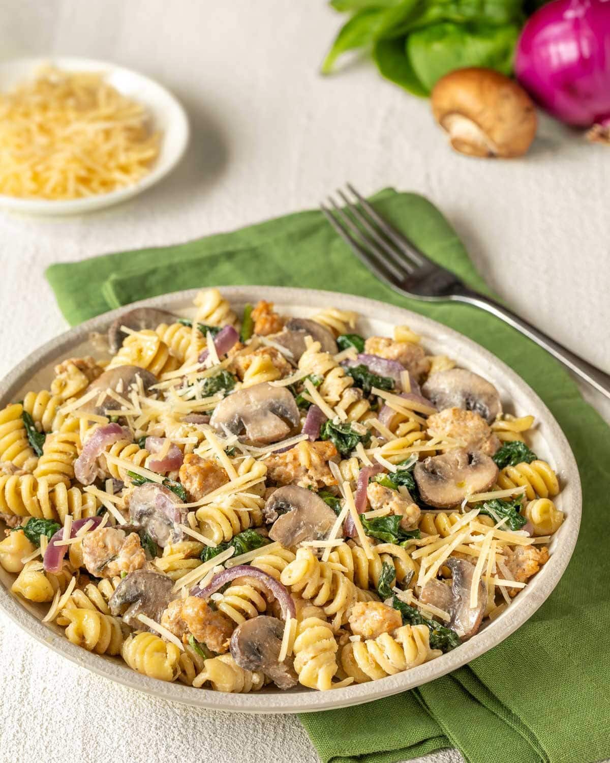 bowl holding a large portion of chicken sausage, spinach and mushroom pasta, with grated Parmesan, a mushroom, and a red onion in the background