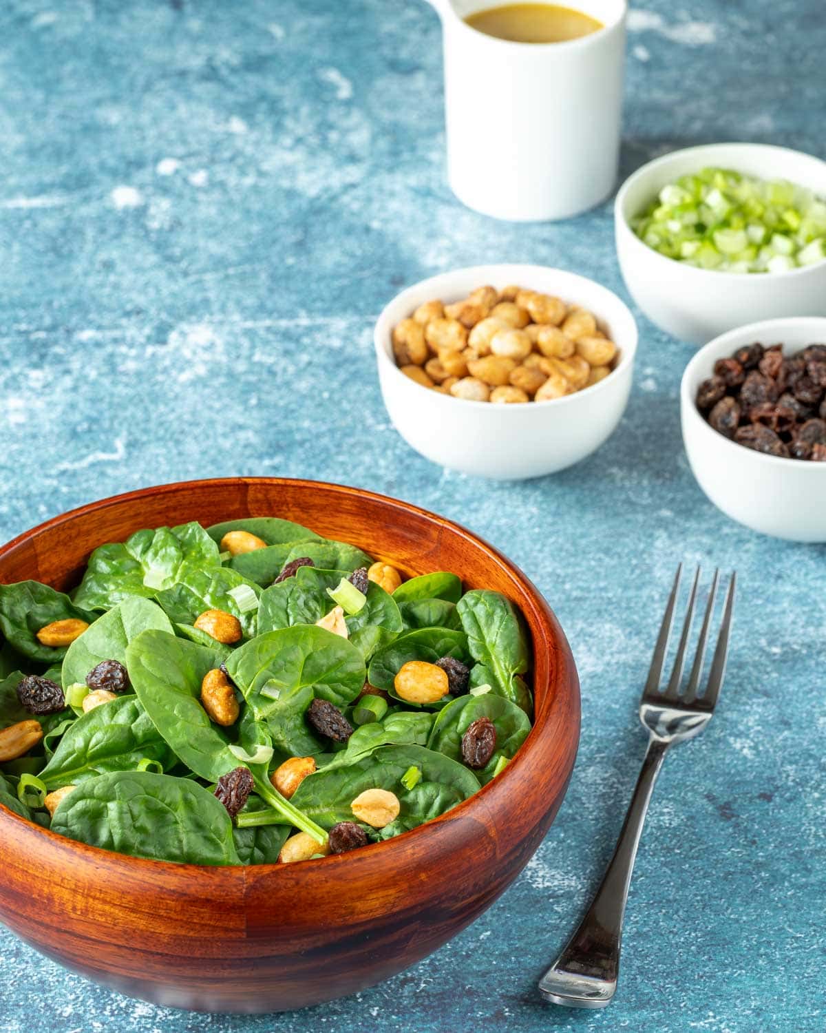 wooden bowl holding a serving of spinach salad with raisins, peanuts and curry dressing on the side