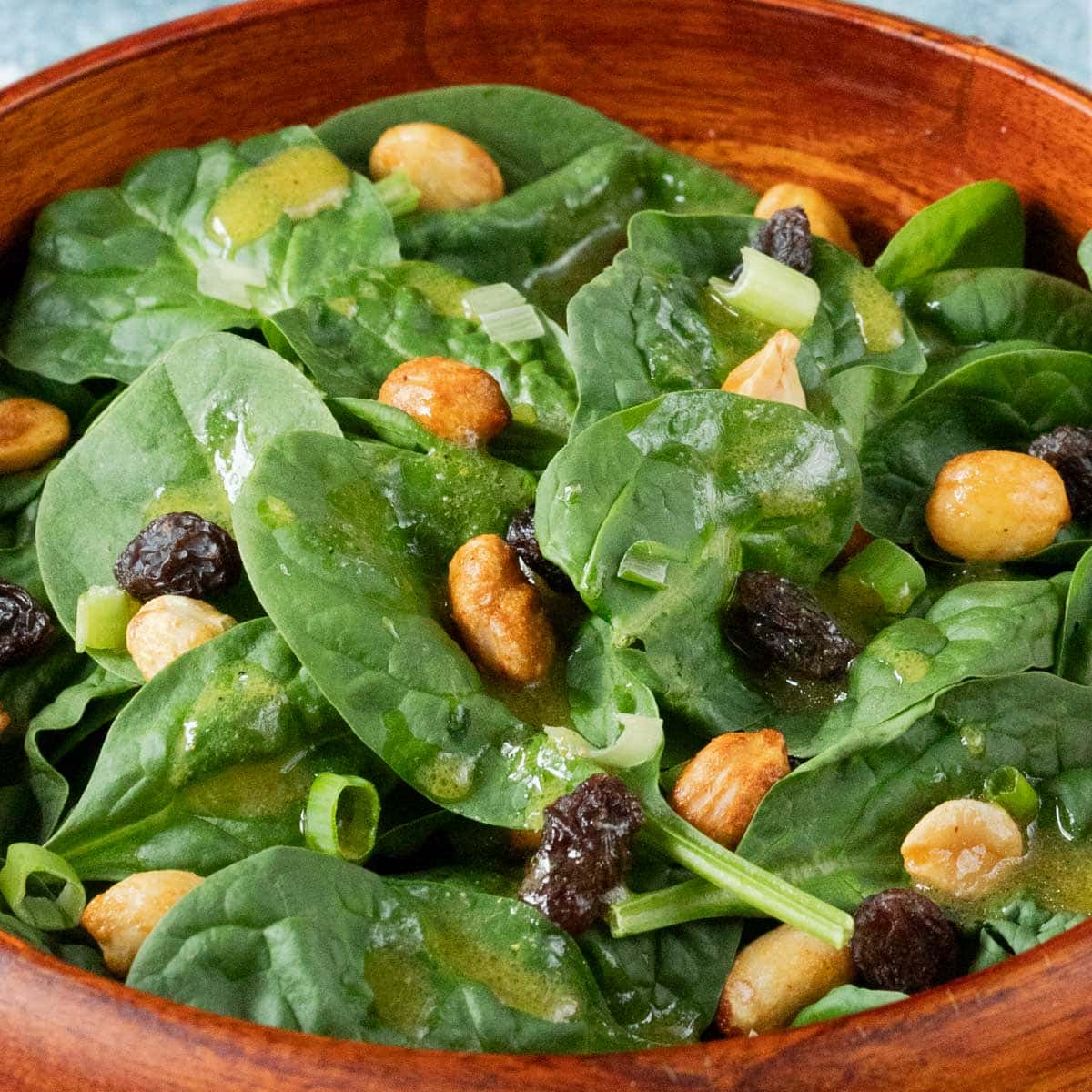 wooden bowl holding a serving of spinach salad with raisins and peanuts and dressing on the side