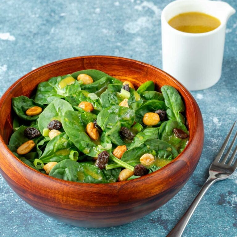wooden bowl holding a serving of spinach salad with raisins and peanuts and dressing on the side