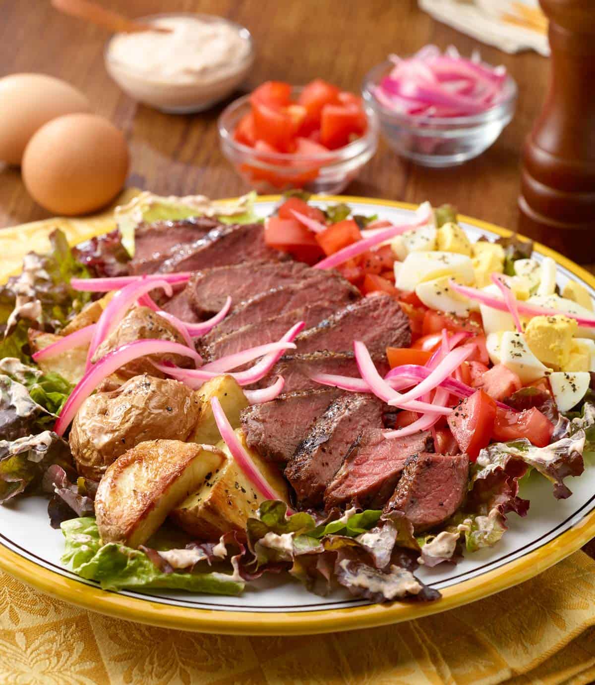 Rows of grilled steak, roasted potatoes, chopped tomatoes and eggs over lettuce