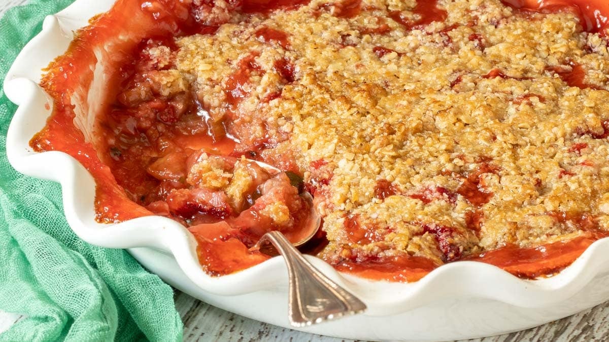 Close up view of Strawberry Rhubarb Crisp in a white pie plate with a scoop removed, and a spoon in the plate