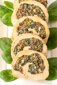Five slices of stuffed pork tenderloin on a white platter, garnished with baby spinach on the side