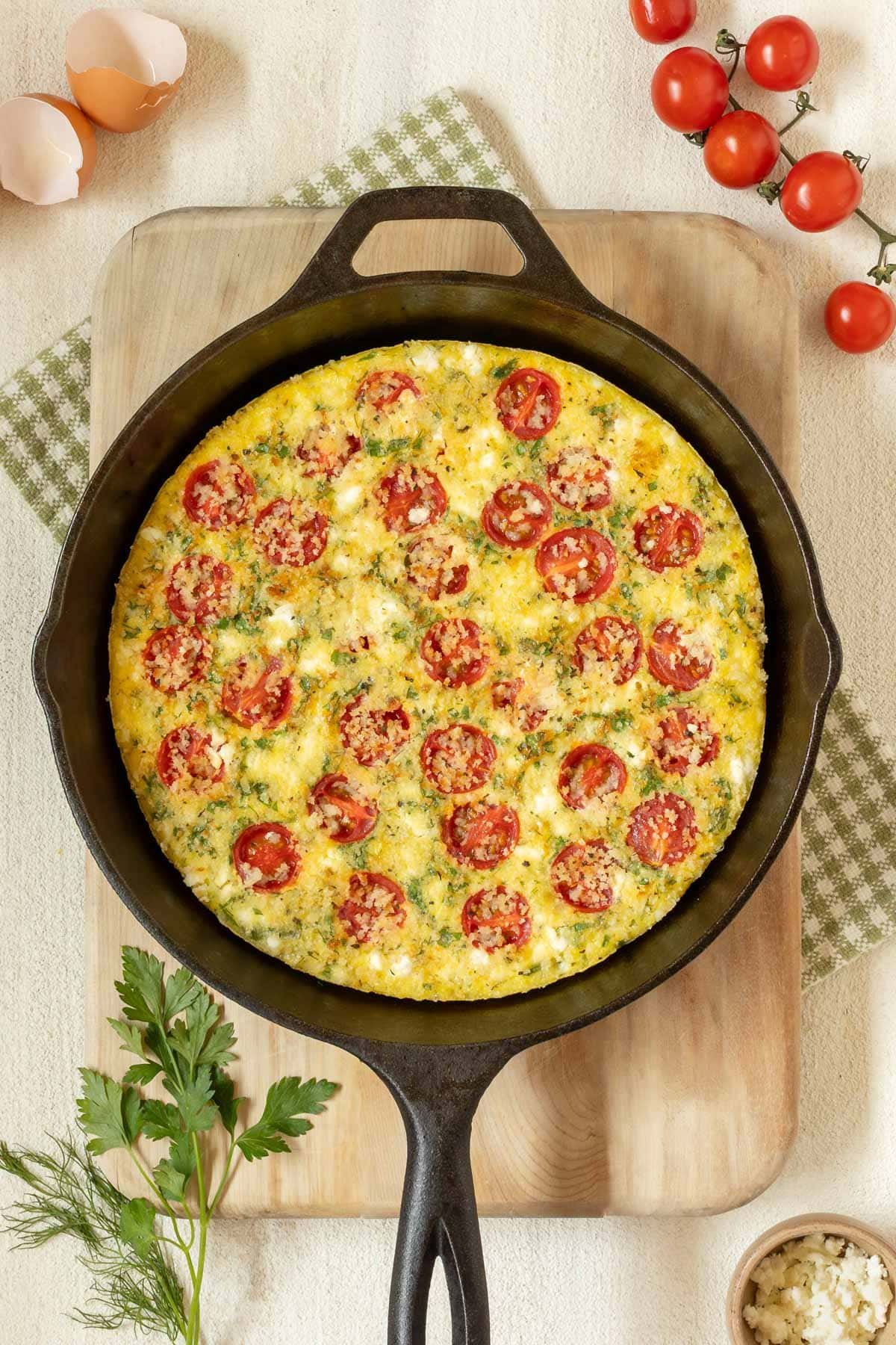 cast iron skillet holding a Tomato Frittata with Feta, on top of a cutting board and surrounded by cherry tomatoes, fresh parsley and dill, feta in a small bowl, and egg shells