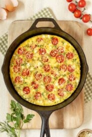 cast iron skillet holding a Tomato Frittata with Feta, on top of a cutting board and surrounded by cherry tomatoes, fresh parsley and dill, feta in a small bowl, and egg shells