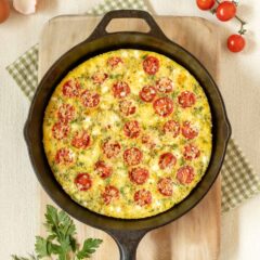 cast iron skillet holding a Tomato Frittata with Feta, on top of a cutting board and surrounded by cherry tomatoes, fresh parsley and dill, feta in a small bowl, and egg shells