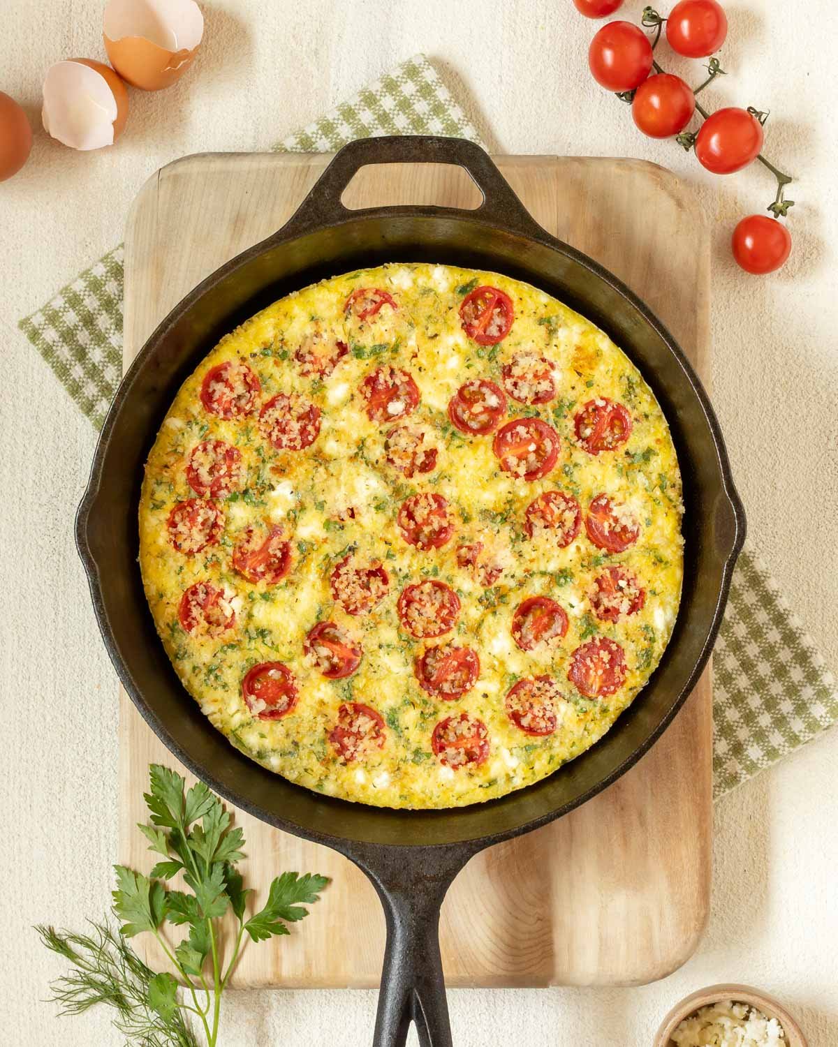 cast iron skillet holding a Tomato Frittata with Feta, on top of a cutting board and surrounded by cherry tomatoes, fresh parsley and dill, feta in a small bowl, and egg shells