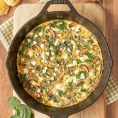 Overhead view of Vegetable Frittata in cast iron skillet set on a cutting board, with mushrooms, cheese, spinach and biscuits around the side